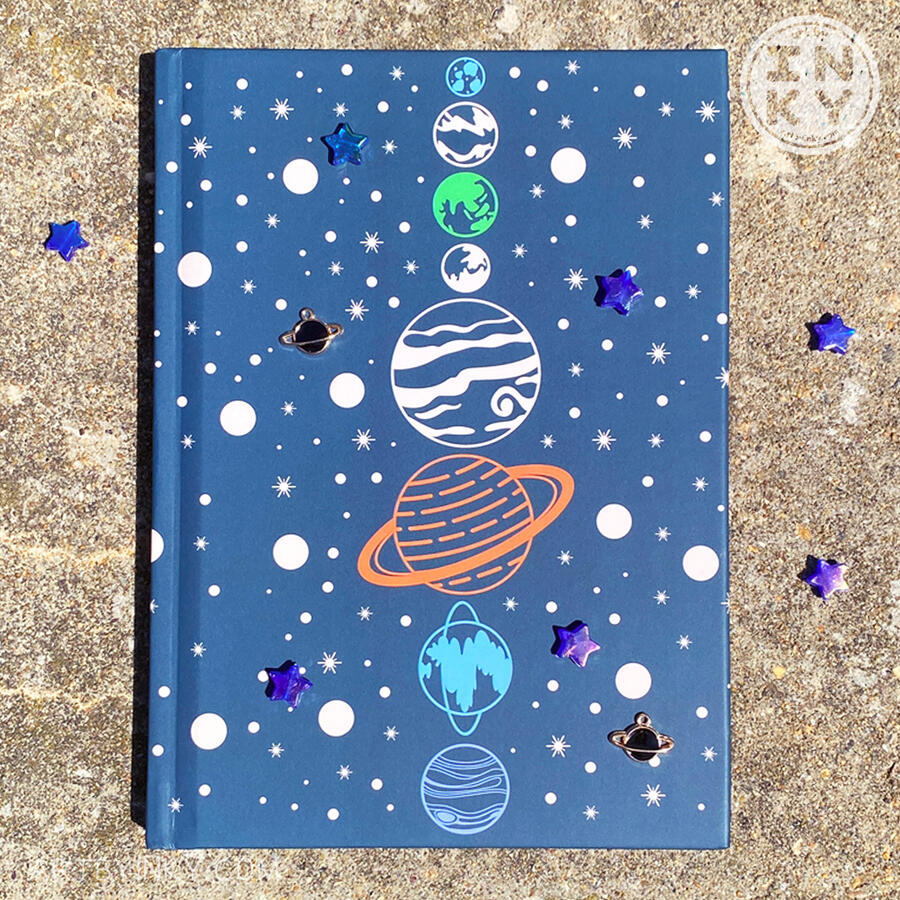 Blue Space Journal A hardcover blue space journal laid flat frontside up with star and planet charms against a cement background outside.