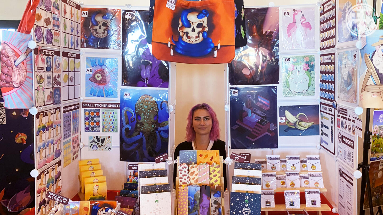 Full front view of the booth layout at an anime convention, surrounded by products like art prints, stickers, washi tape tote bags, journals, bookmarks, pins, jewelry, and coin purses.