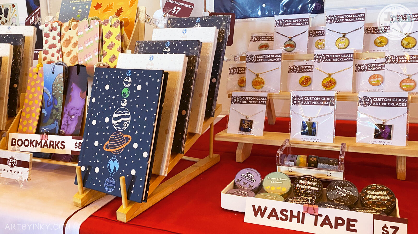 Close up of the booth layout at an anime convention showing products like journals, bookmarks, jewelry, and washi tape.